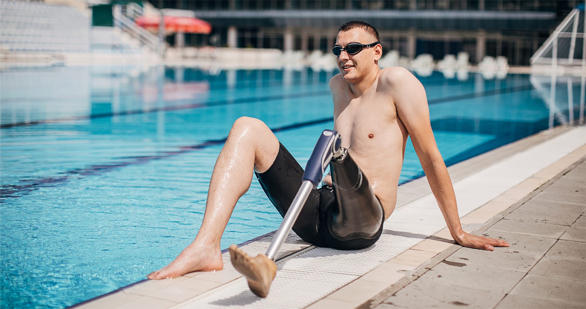 Man with a prosthetic leg, wearing a bathing suit and swim goggles sitting near the edge of a pool 