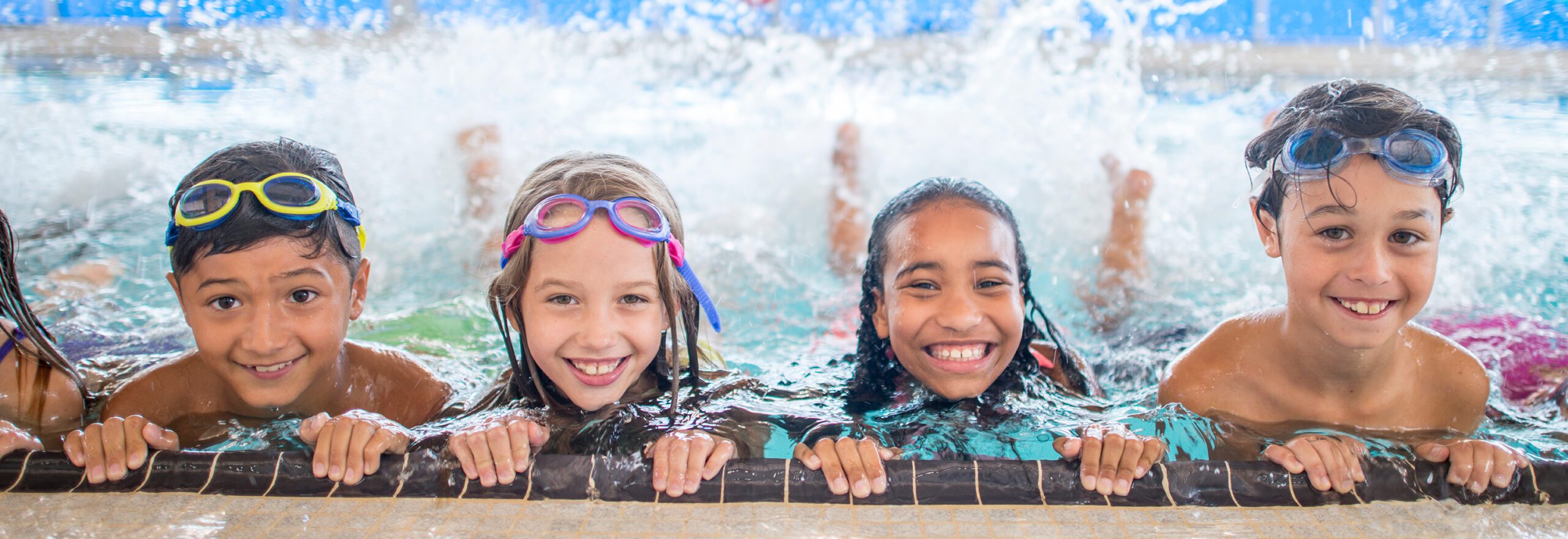 Four children in pool, holding pool edge and kicking, smiling at camera